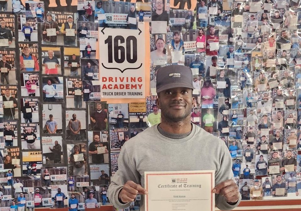 Man wearing blue jeans, grey shit, and cap...standing in front of photos of graduates at 160 Driving Academy in Rockford IL.