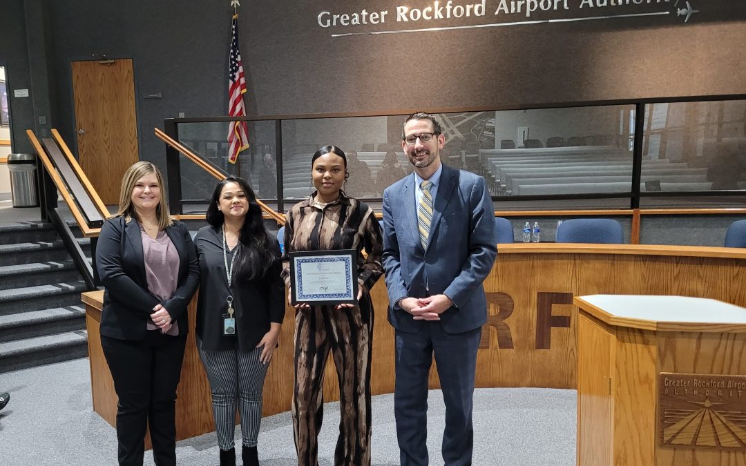 4 people standing in front of a large meeting desk. The 2 women on the right are staff members of The Workforce Connection, the 3rd woman (L-R) is Alyssa who overcame barriers to complete her training. She is holding the award. On the right is City of Rockford's Mayor Thomas McNamara